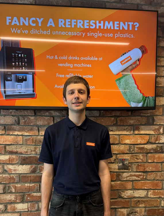Smiling easyHotel employee in a branded navy polo shirt, standing in an easyHotel lobby with a brick wall background. Behind him, a digital screen promotes easyHotel's eco-friendly refreshments, vending machines, and free refillable water.