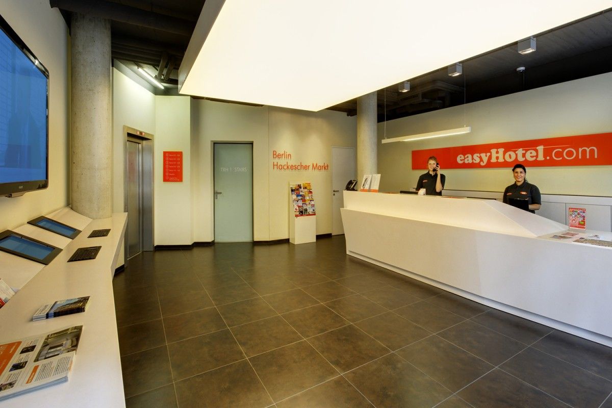 easyHotel Berlin Hackescher Markt lobby featuring a white reception desk with two staff members, a multi-screen information kiosk, and a prominent easyHotel.com sign.