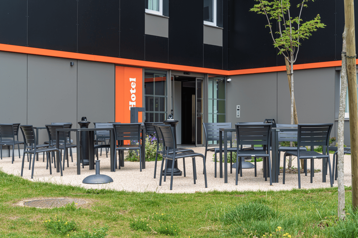 Exterior view of the easyHotel CDG, showcasing its modern black and grey facade with a distinctive orange stripe and vertical 'Hotel' sign. An inviting outdoor patio area with dark grey metal tables and chairs is set on gravel, bordered by green grass and