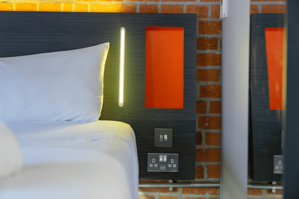 A modern, compact easyHotel Manchester room showing a bed with white bedding, a dark grey headboard featuring an integrated vertical light, and a signature orange panel. Below the light, UK power outlets with a USB port are visible against an exposed bric