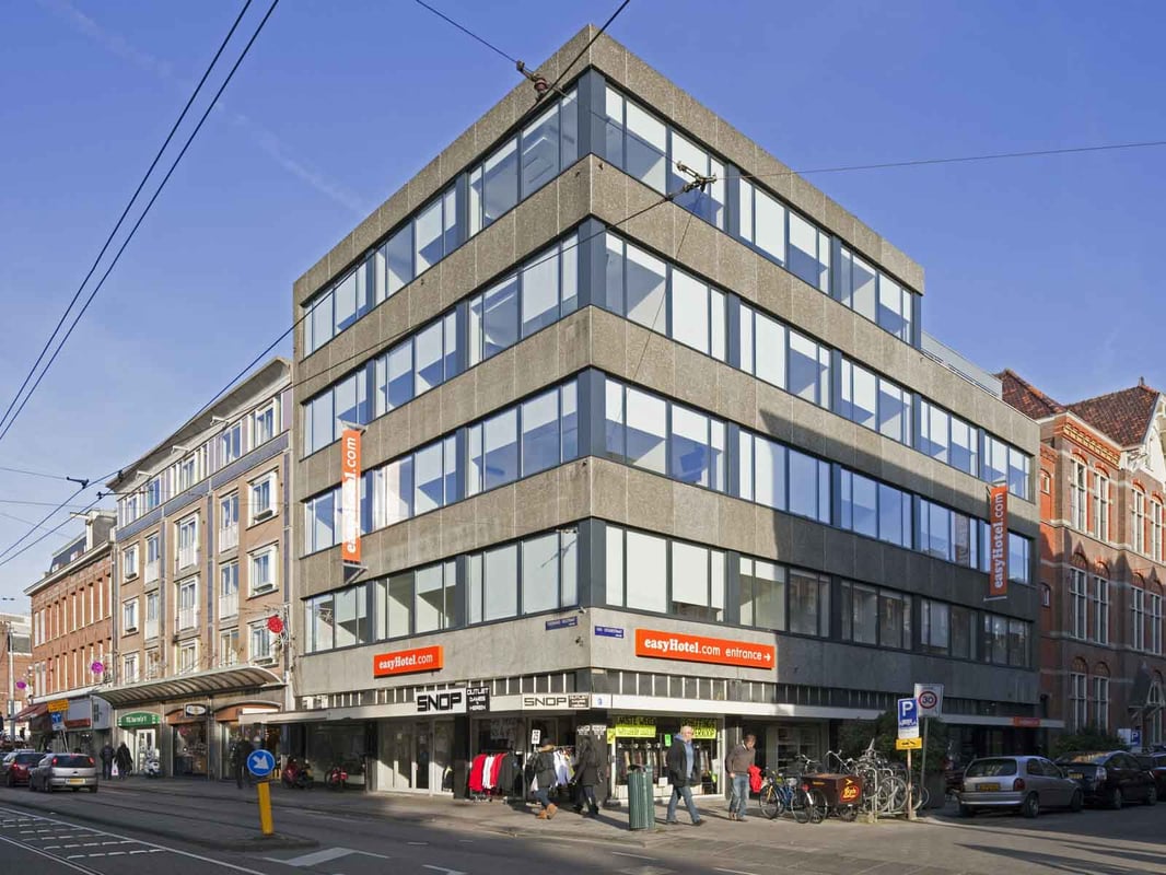 Modern dark easyHotel Amsterdam City Centre South building at night, with illuminated windows, glowing streetlights, and red light trails from traffic on a busy urban street.