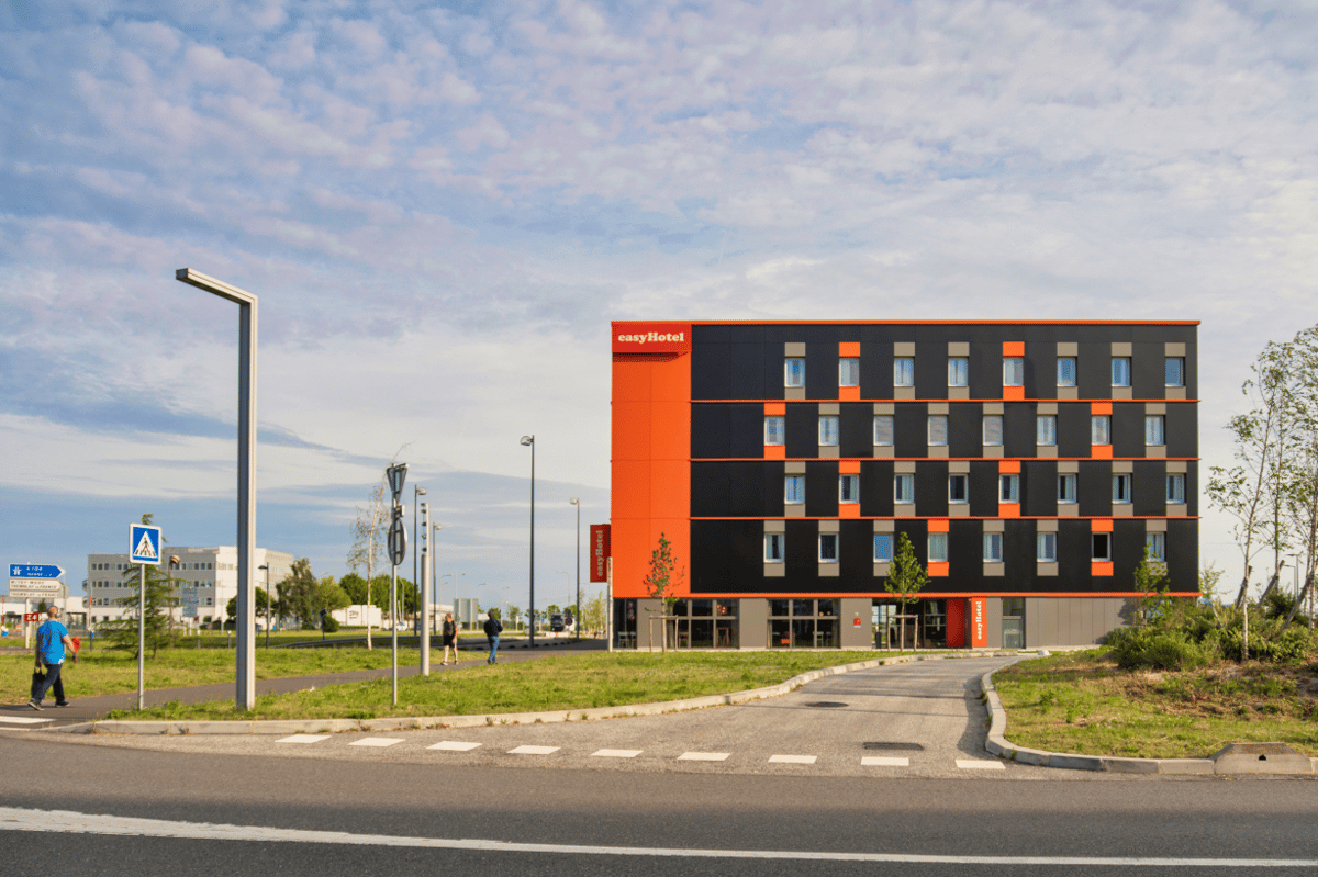 Modern easyHotel building exterior at Charles de Gaulle Airport (CDG), featuring its distinctive orange and dark gray facade and surrounding access roads.