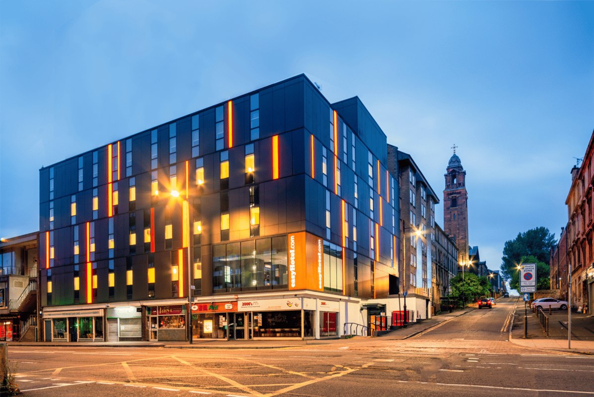 Modern easyHotel Glasgow building exterior at twilight, featuring its distinctive dark facade with vertical orange lighting strips and illuminated windows, prominent easyHotel.com branding, and a historic clock tower in the background.