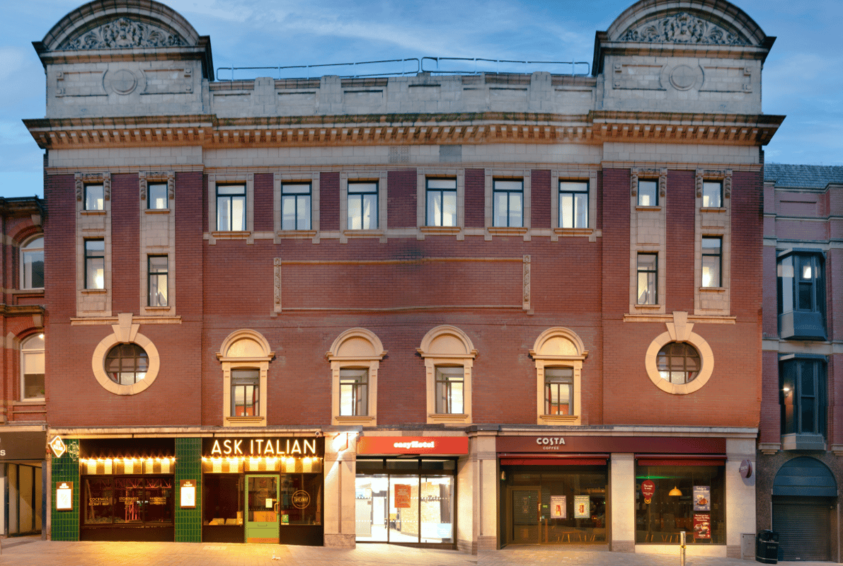 Facade of easyHotel Leeds, a prominent red brick building with ornate light stone architectural details and multiple windows. The ground floor features entrances for easyHotel, ASK ITALIAN, and Costa Coffee.