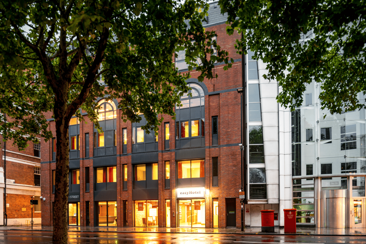 Exterior view of the easyHotel London City Shoreditch building at dusk, featuring a red brick facade, illuminated windows, and the lit easyHotel sign. A large tree with green leaves is in the foreground, and the wet street reflects the warm interior light