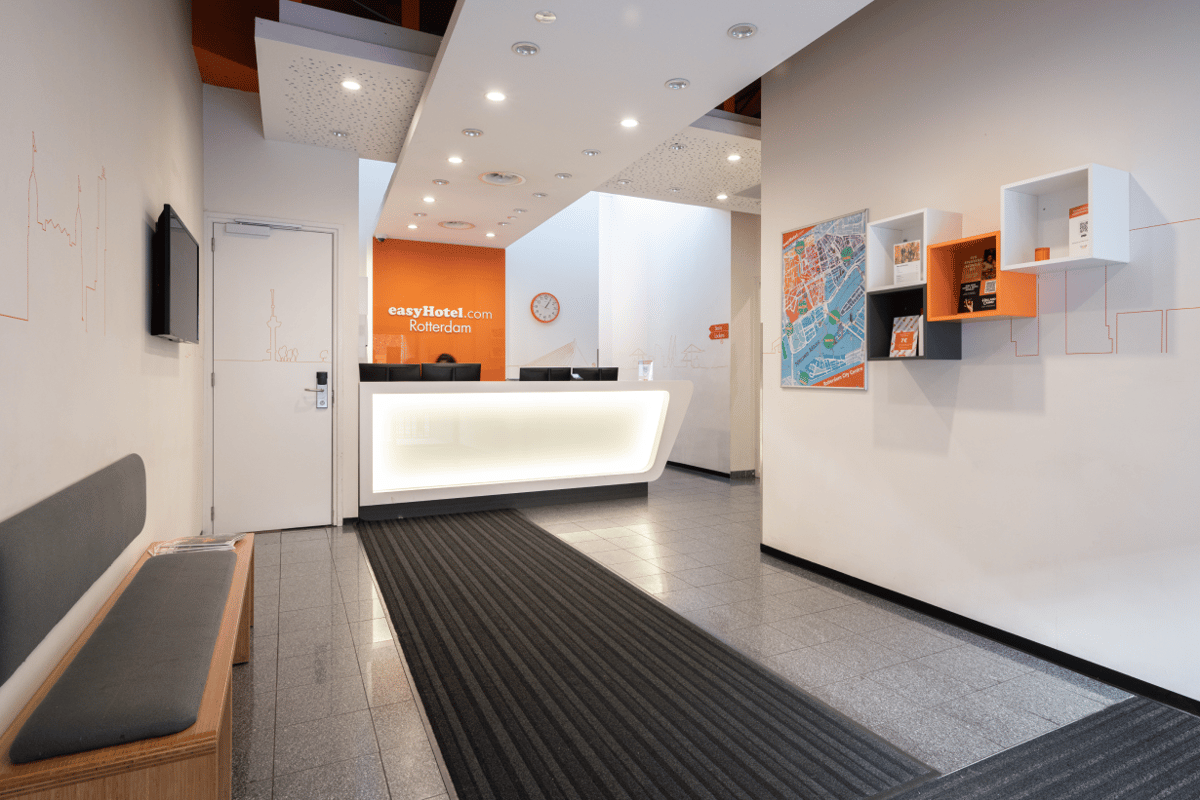 Modern and brightly lit easyHotel Rotterdam City Centre lobby featuring an illuminated reception desk, orange branding, and local city map with skyline illustrations.