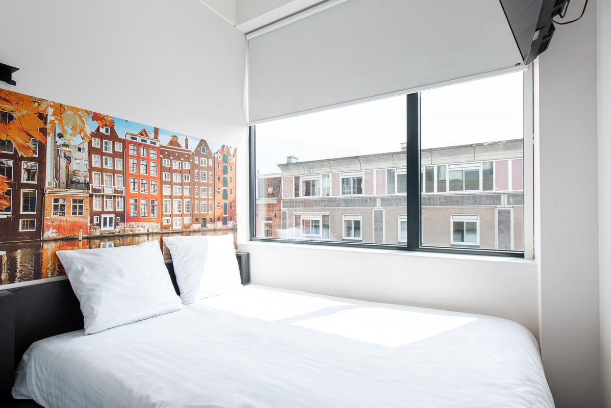 An easyHotel room in Amsterdam City Centre South featuring a brightly lit bed with white linen. Behind the bed is a large mural depicting colorful traditional Dutch canal houses along a canal. A wide window provides a view of a modern building across the