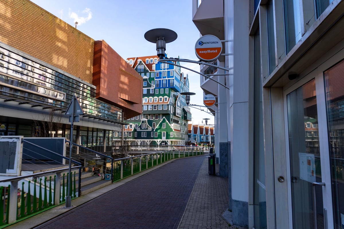 Exterior view of an easyHotel building with its prominent orange circular sign, situated on a paved street in Zaandam, Amsterdam, with a large building featuring stacked traditional Dutch house facades in the background.
