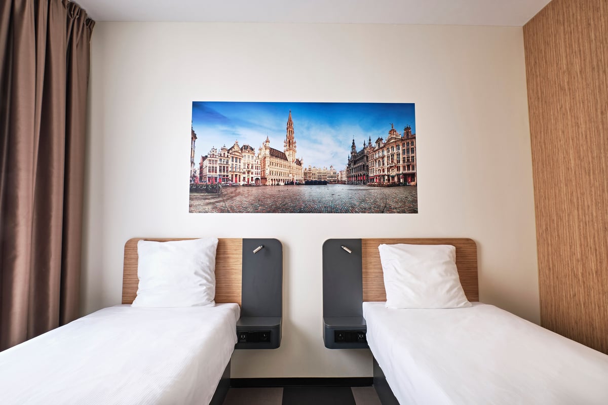 An easyHotel Brussels City Centre room featuring two comfortable single beds with white linens and a large panoramic print of the iconic Grand Place in Brussels displayed above them.