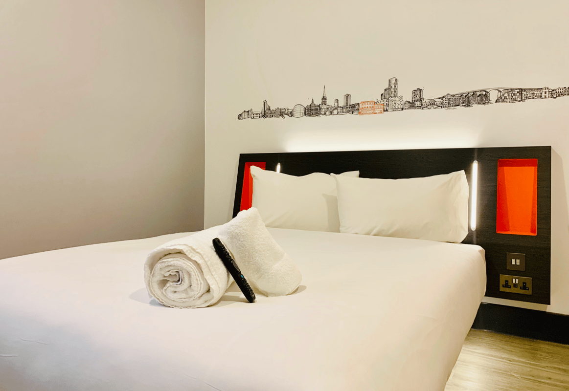 easyHotel Ipswich room with a king-size bed, crisp white linen, and rolled towels. The modern black headboard features orange accents, integrated lighting, and UK power outlets. A unique black line-art cityscape mural of Ipswich landmarks with subtle oran