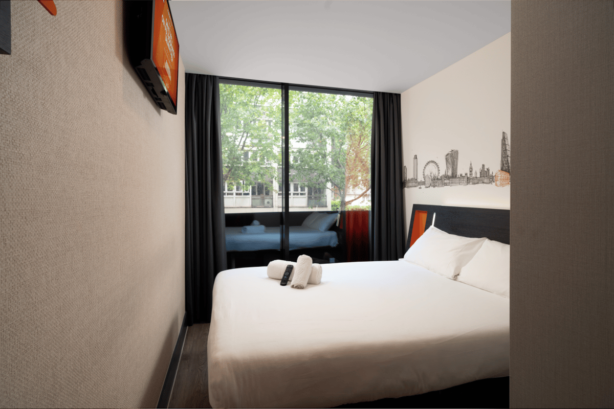 A bright, minimalist easyHotel room in London City Shoreditch, featuring a double bed with white linen, a wall-mounted TV, a black and orange headboard, and a London skyline mural above the bed. Large windows offer a view of green trees outside.