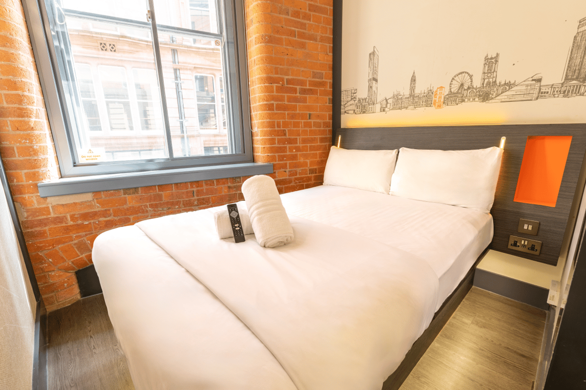 An easyHotel Manchester room featuring a double bed with white linen, a distinctive Manchester city skyline mural above the headboard, and an exposed brick wall.