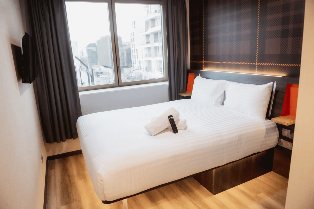 A modern easyHotel room in Paris Nord Aubervilliers featuring a crisp white double bed, plaid accent wall, wall-mounted TV, and a window overlooking city buildings.