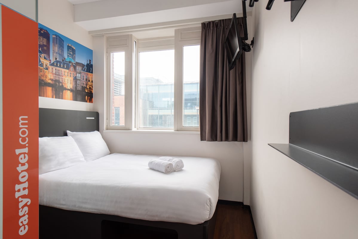 A bright, compact easyHotel room in The Hague, featuring a bed with crisp white linen, a large window, a wall-mounted TV, and a striking mural of the city's skyline above the bed.