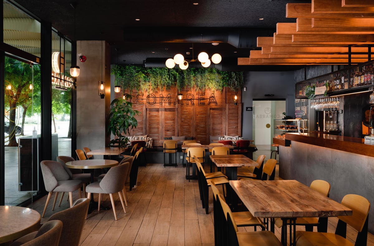 Modern cafe and bar interior in Barcelona, featuring wooden tables, cushioned chairs, and a central bar area. The warm-lit space includes a green plant wall above wooden panels with "COMAREA" lettering, alongside large windows overlooking trees.