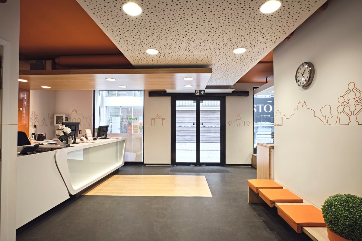 ALT: Modern easyHotel Brussels City Centre lobby featuring a white reception desk, orange accented ceiling and seating, and outline art of Brussels landmarks on the walls.