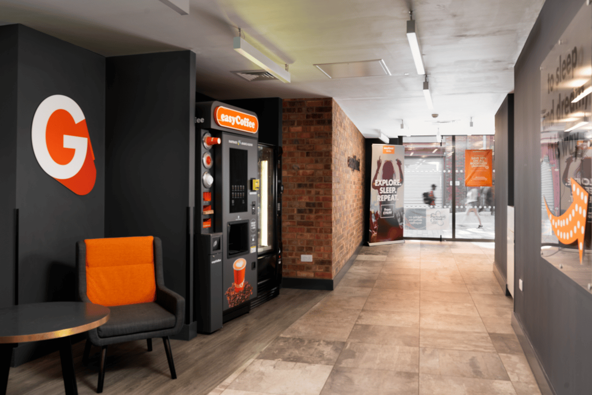 Lobby area at easyHotel Leeds with an easyCoffee vending machine, a gray chair with an orange backrest, a small table, a brick accent wall, and an "EXPLORE. SLEEP. REPEAT." banner.