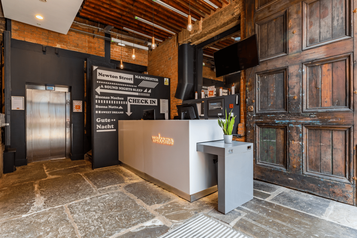 easyHotel Manchester lobby featuring a modern white reception desk with a glowing "welcome" sign, exposed brick walls, a wooden ceiling, and a black wall with white text directions including "Check In" and "Manchester." A silver elevator door and rustic w