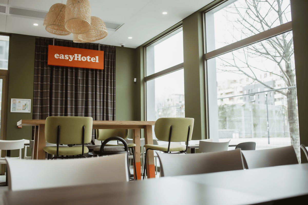 Interior view of the easyHotel Paris Nord Aubervilliers common area, featuring modern tables and chairs in a brightly lit space with large windows. The easyHotel logo is prominently displayed on an orange sign.