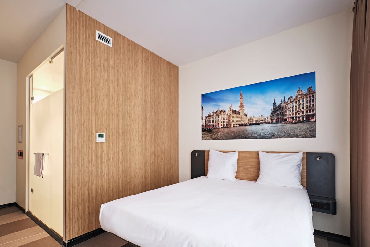 An easyHotel room in Brussels City Centre featuring a double bed with white linen, a light wood headboard, and built-in bedside shelves. Above the bed, a large panoramic print of Brussels' iconic Grand Place (Grote Markt) is prominently displayed. An inte