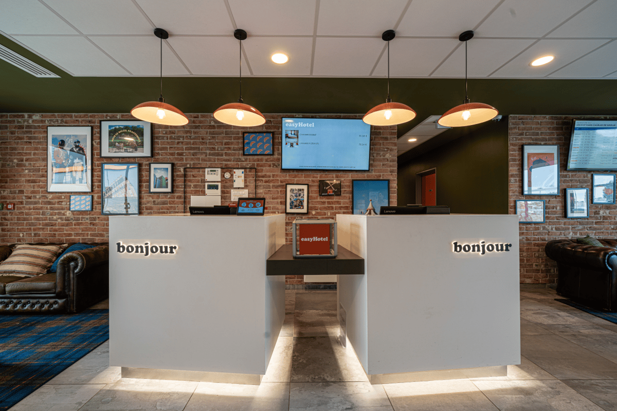 easyHotel CDG Airport lobby reception area with white desks, 'bonjour' signs, and a brick wall decorated with art.