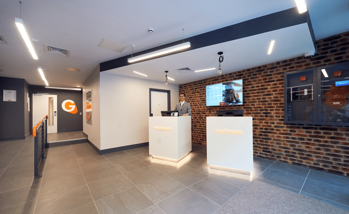 Modern reception lobby of easyHotel London City Shoreditch, featuring a friendly staff member, two glowing 'welcome' self-check-in kiosks, a stylish exposed brick wall, and a large screen display.