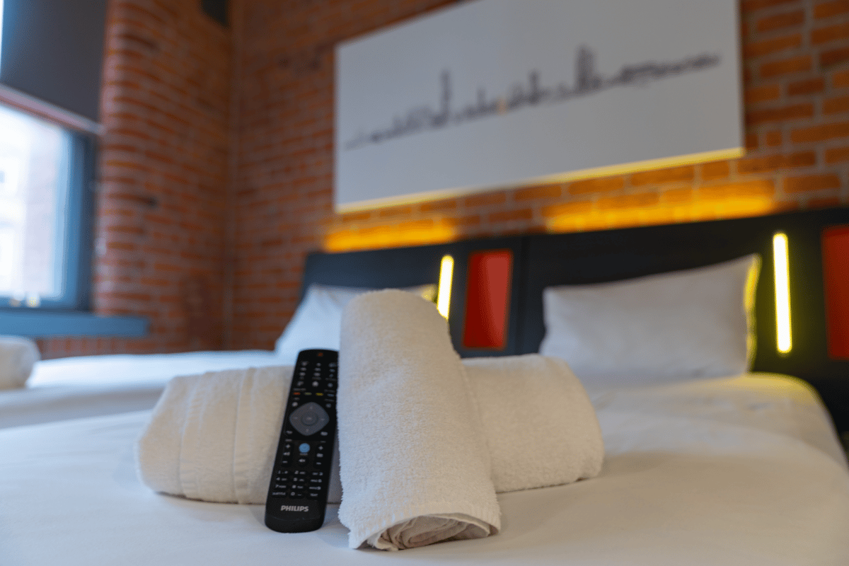 A clean easyHotel Manchester room featuring fresh white towels and a TV remote on a pristine white bed, with an exposed brick wall and a modern headboard with accent lighting in the background.