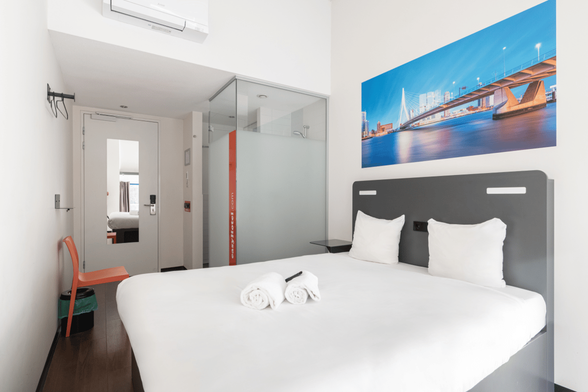 A modern easyHotel room in Rotterdam City Centre featuring a white bed, orange chair, dark wood floor, and a large wall artwork depicting the iconic Erasmus Bridge.