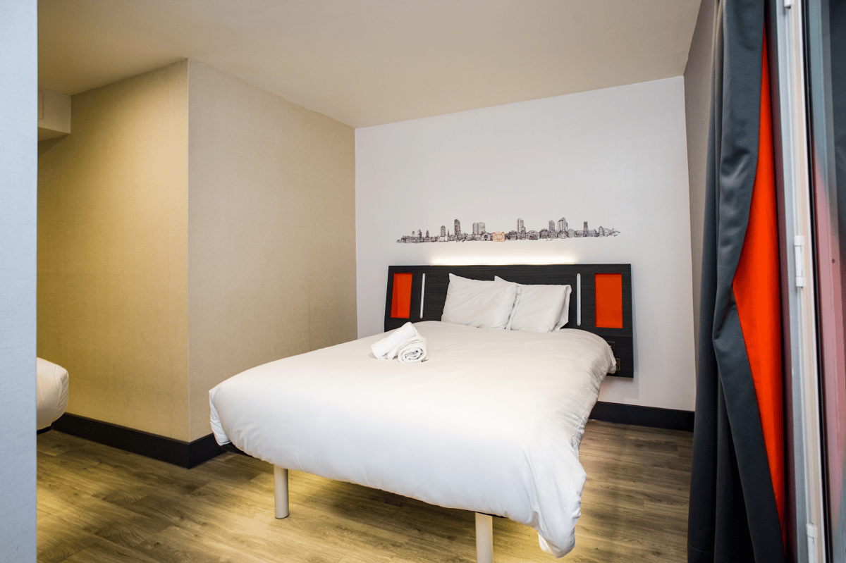 easyHotel Leeds double room with minimalist design, featuring a bed with white bedding, black and orange headboard, a cityscape mural on the wall, and wood-effect flooring.