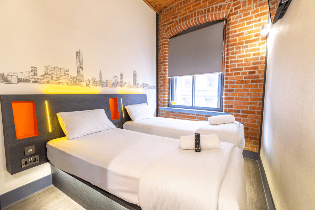 Modern easyHotel Manchester twin room with white beds, a dark headboard with orange accents and yellow lighting, a Manchester city skyline mural, and an exposed brick wall.