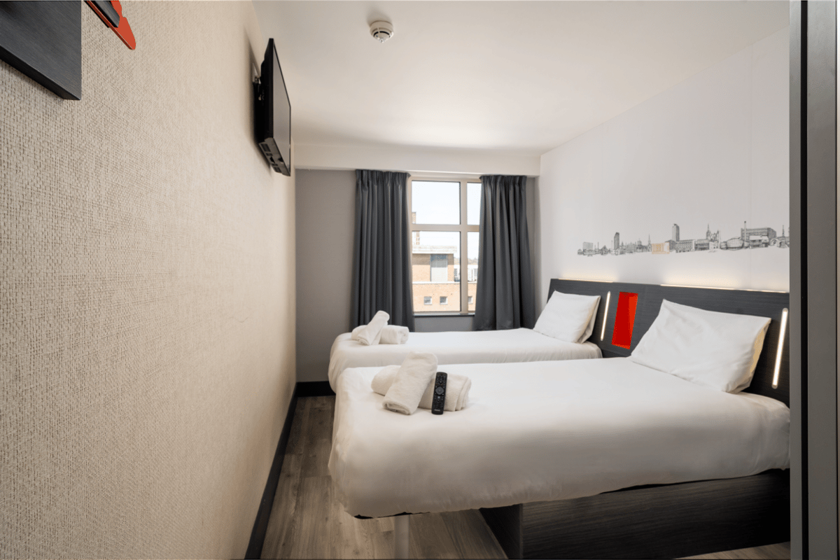 easyHotel Sheffield twin room with two white beds, a wall-mounted TV, and a Sheffield cityscape mural above the dark grey headboards.
