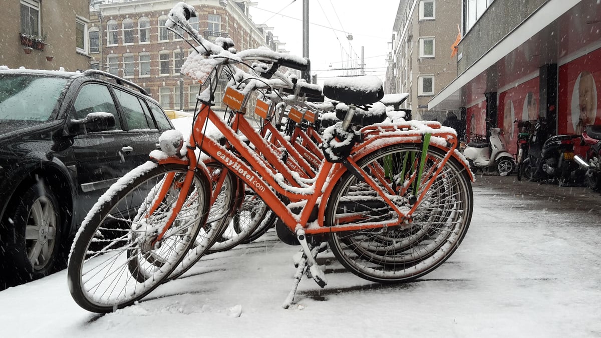 Row of orange easyHotel bicycles heavily covered in snow, parked on a snowy street in Amsterdam City Centre South, with buildings and a black car in the background.