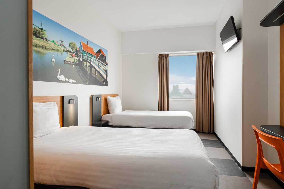 A clean and bright easyHotel twin room in Amsterdam Zaandam, featuring two single beds with white striped bedding, a large wall art depicting a scenic Dutch canal with windmills and swans, a mounted TV, a functional desk with an orange chair, and a window
