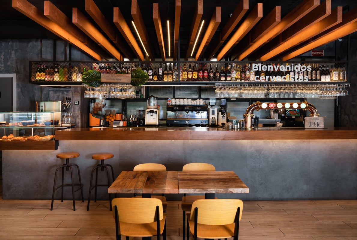 Modern bar and cafe area inside easyHotel Barcelona, featuring a wooden counter, beer taps, espresso machine, pastry display, and a "Bienvenidos Cerveceristas" sign.