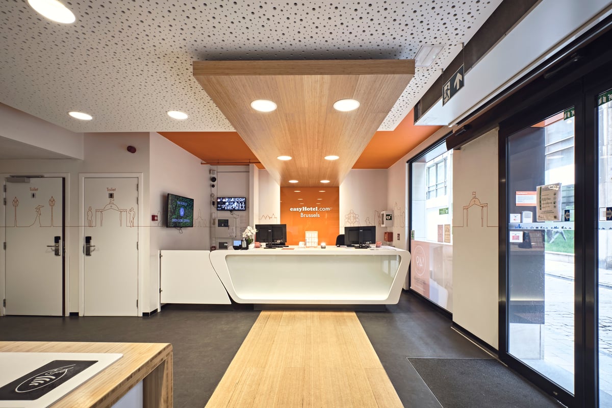 A bright, modern reception lobby at easyHotel Brussels City Centre. The space features a sleek white reception desk, a prominent orange accent wall with the easyHotel logo, a light wood ceiling panel, and a matching light wood floor path leading towards t