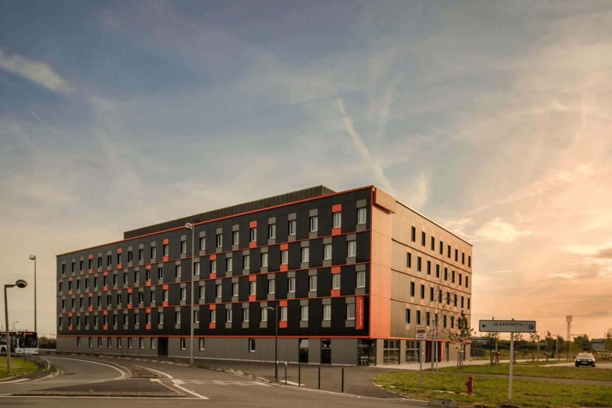 Exterior of the easyHotel CDG building with its distinctive modern dark grey and orange facade, seen from street level at sunset with a warm sky.