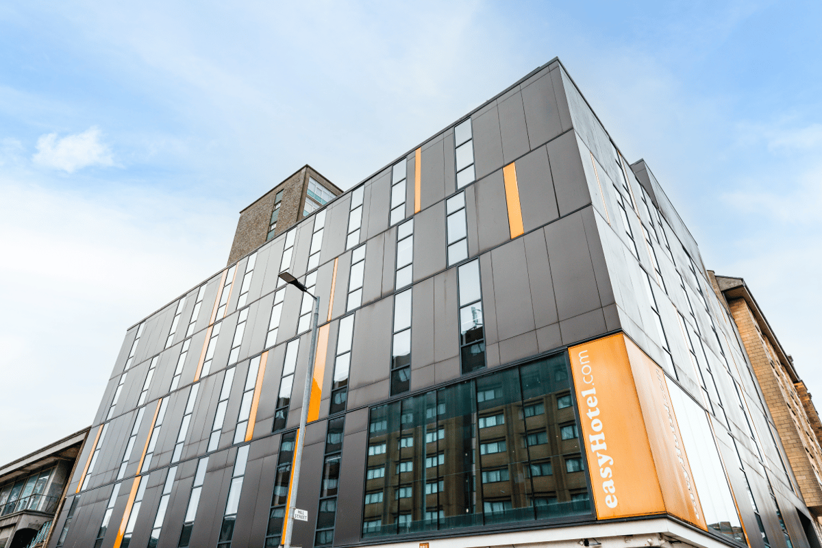 Exterior view of the easyHotel Glasgow building, a modern structure with a dark grey facade, striking orange vertical panels, and many windows, under a clear blue sky.