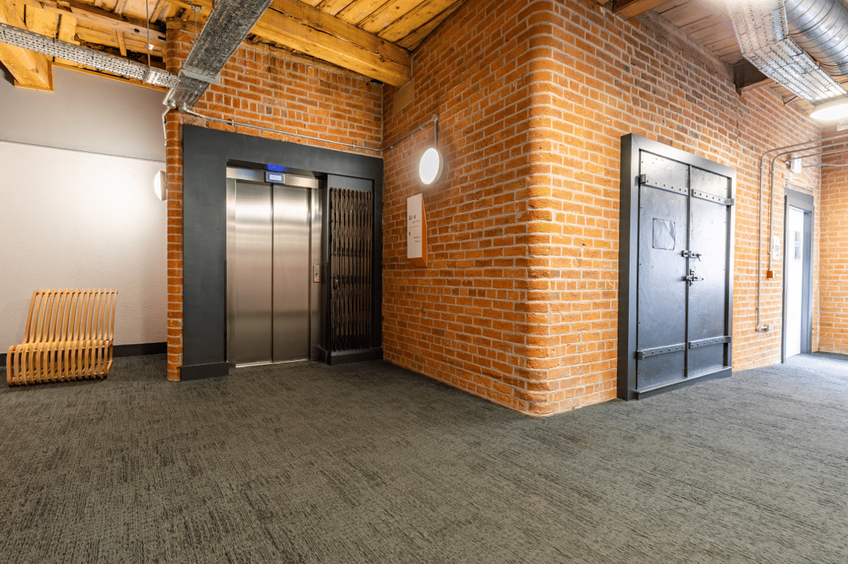 Modern industrial-style lobby at easyHotel Manchester, featuring exposed red brick walls, a natural wood ceiling, a sleek stainless steel elevator, and a unique light wood bench on dark grey patterned carpet.