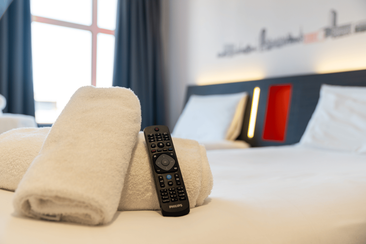 Clean white towels and a TV remote control on a bed in a modern easyHotel room in Sheffield, featuring a dark headboard with red and yellow lighting details.