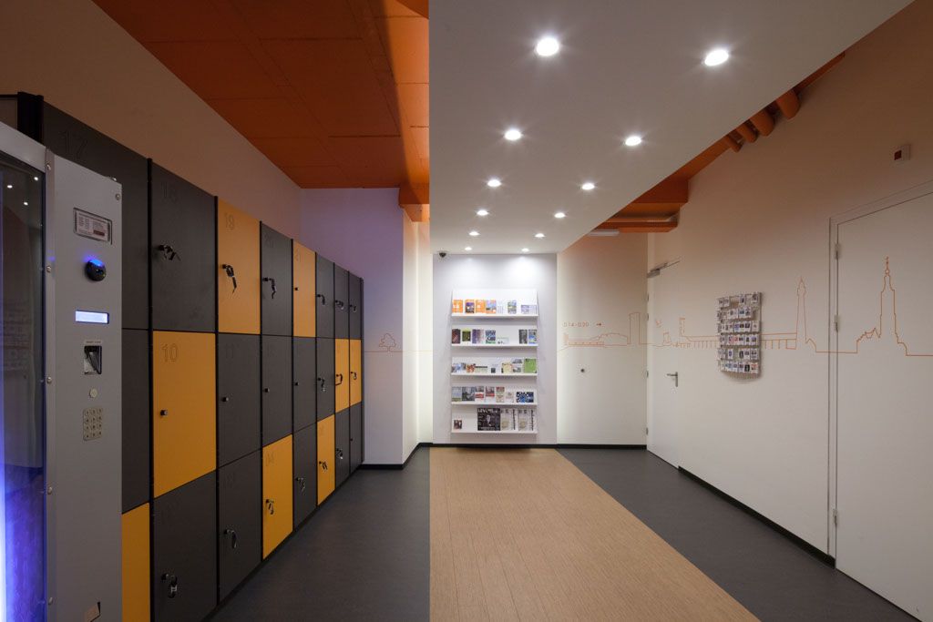 A brightly lit modern hallway at easyHotel The Hague featuring rows of black and yellow guest lockers, a self-service machine, a white wall-mounted brochure display, and an orange skyline mural on the white wall.