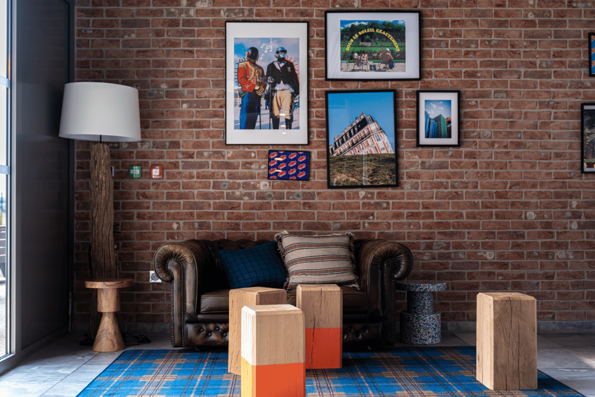 EasyHotel Paris CDG lounge featuring a brown leather Chesterfield sofa, exposed brick wall with eclectic art, and a blue plaid rug. The space includes a tree trunk floor lamp and wooden decorative blocks, creating a comfortable and modern aesthetic.