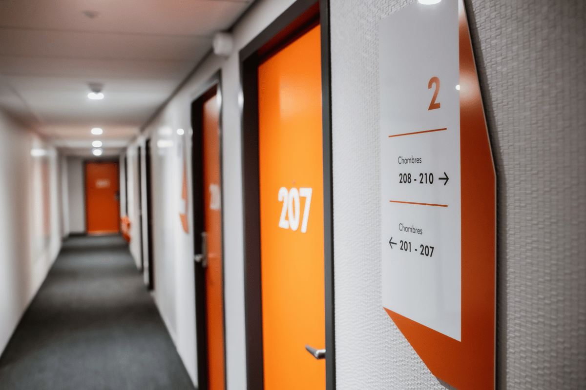 A modern, well-lit hotel hallway with vibrant orange doors leading to guest rooms at easyHotel Paris Nord Aubervilliers. White walls and a grey carpet create a clean, contemporary design, with clear signage indicating room numbers 201-210.