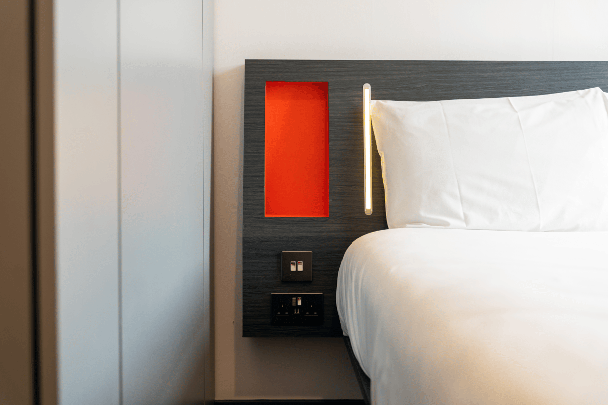 Close-up of an easyHotel Sheffield room featuring a bed with crisp white linens, a dark headboard with a vibrant orange cubby, integrated bedside lighting, and convenient power outlets with USB ports.