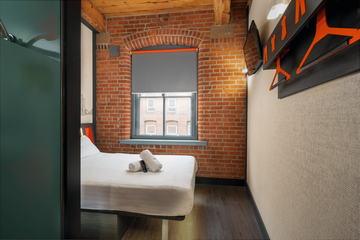 Modern easyHotel Manchester room with a white bed, exposed brick wall, large window with a grey blind, and orange-accented features including a wall-mounted TV and hanging unit, set on a wooden floor.