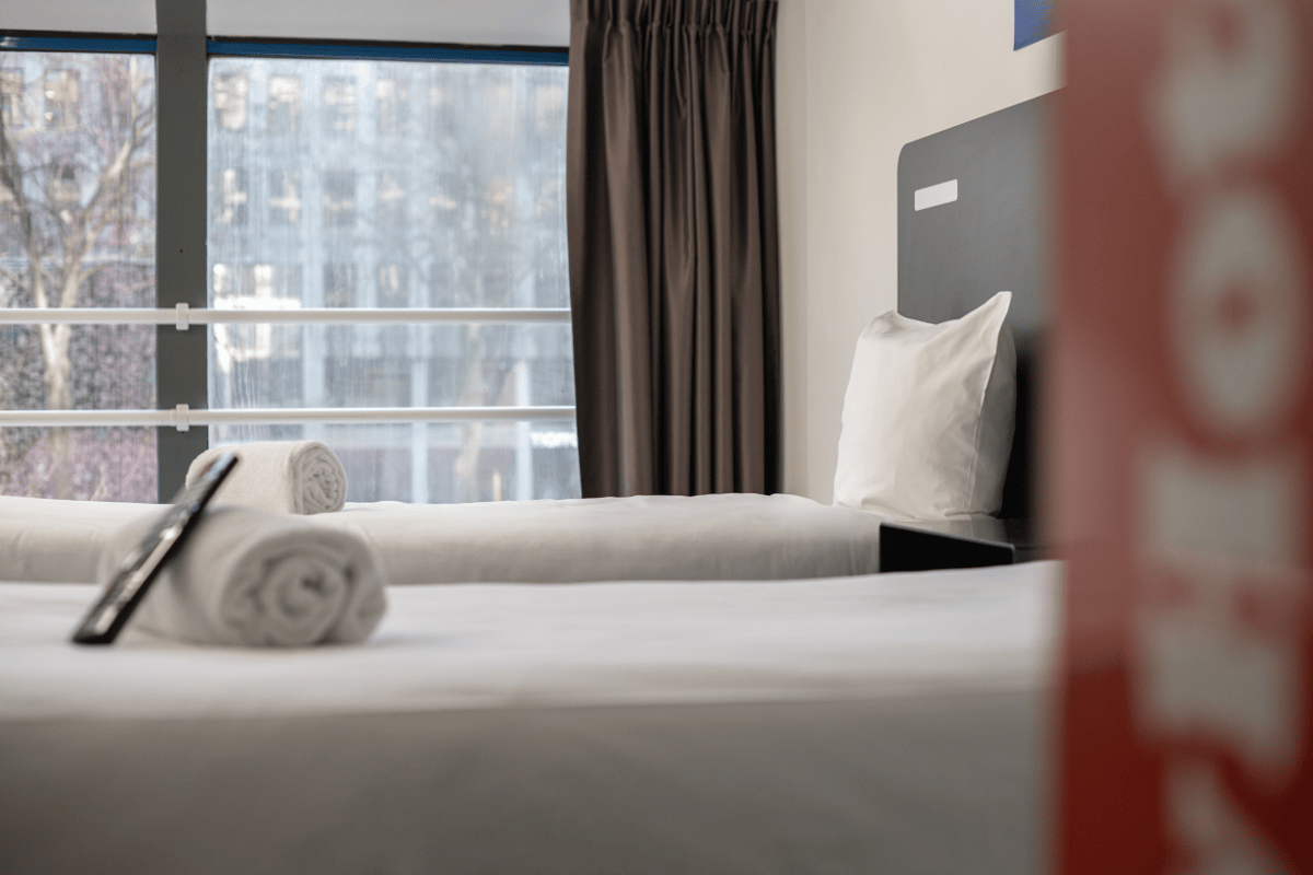 An easyHotel Rotterdam City Centre room featuring two minimalist beds with crisp white linens, a grey headboard, and a large window offering a view of city buildings. Rolled white towels and a TV remote are visible on the bed in the foreground.
