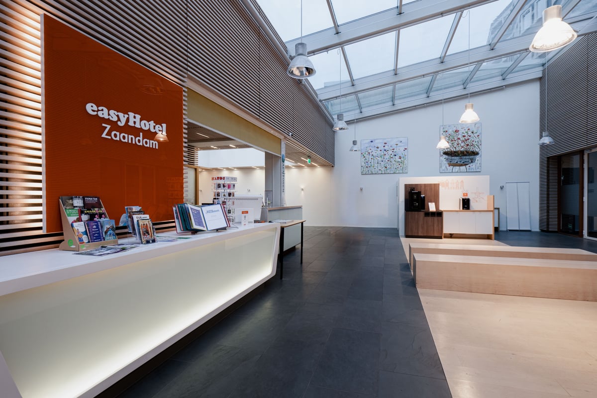 Modern and bright easyHotel Zaandam lobby featuring a white reception desk with an orange easyHotel Zaandam sign, a large skylight overhead, and a self-service coffee station.