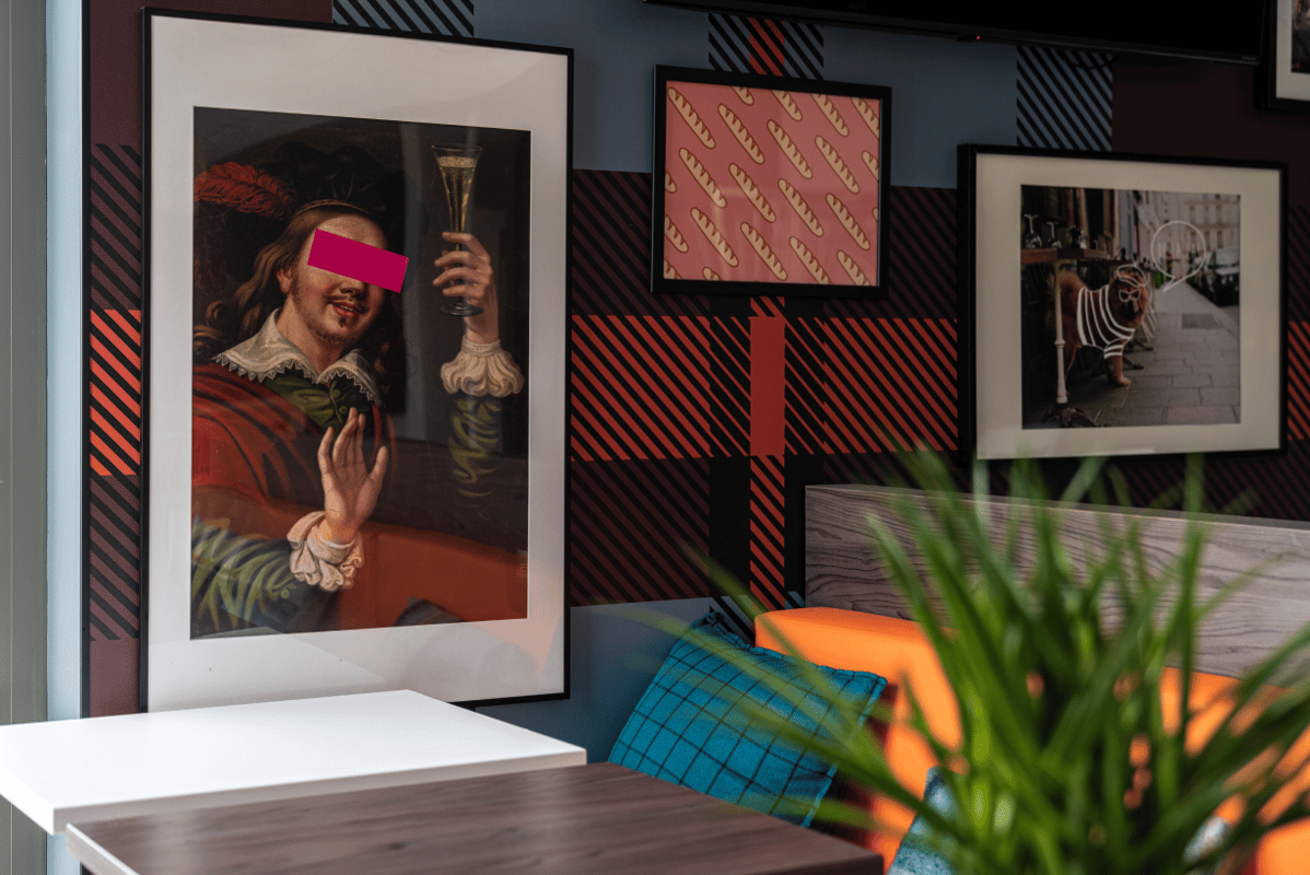 Modern and vibrant easyHotel lounge area, likely easyHotel Paris Charles de Gaulle, featuring a red and black plaid wall. Quirky framed art includes a classical portrait holding champagne, a baguette pattern, and a humorous dog on a Parisian street, compl