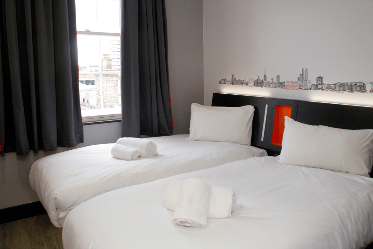 Modern easyHotel Ipswich room featuring two single beds with white bedding and rolled towels. Above the dark headboard, an Ipswich cityscape mural in black, white, and orange. A window with grey curtains overlooks city rooftops.