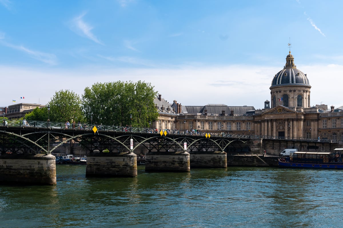 View of the Pont des Arts bridge over the Seine River in Paris, with the historic Institut de France building and its prominent dome on the far bank under a clear blue sky.