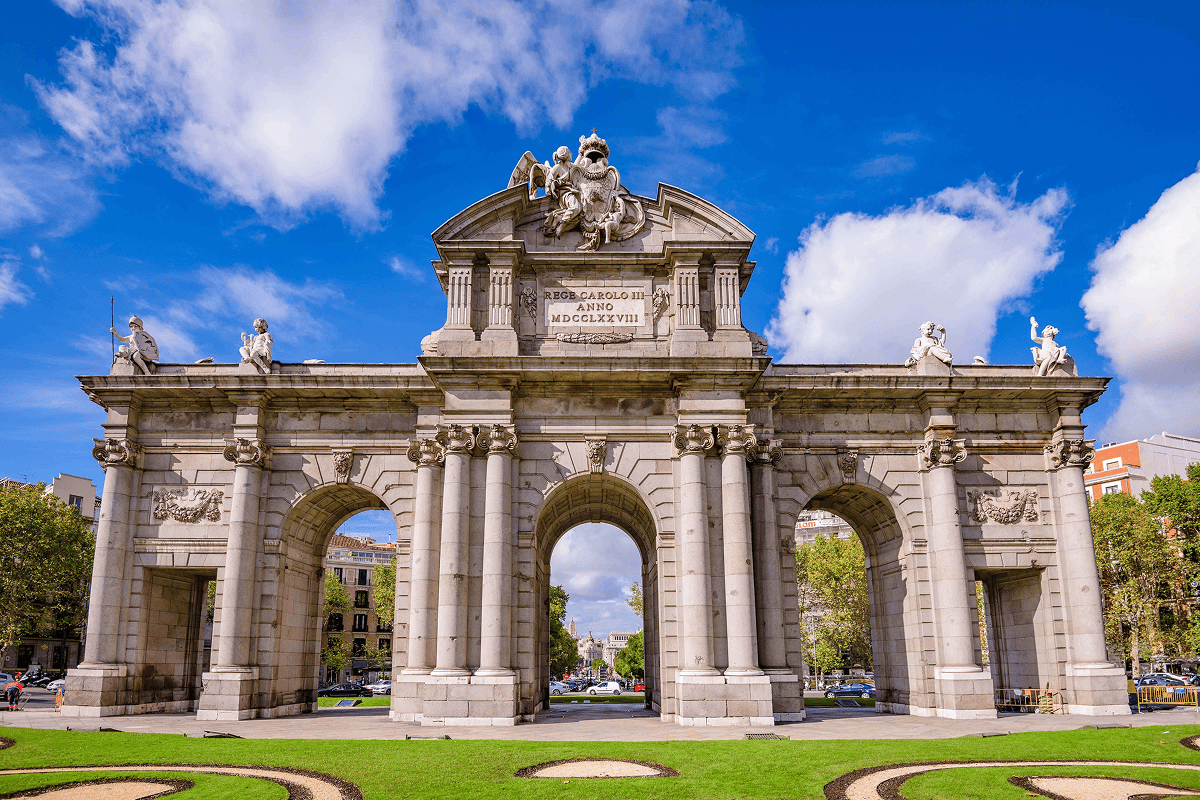 The historic Puerta de Alcalá in Madrid, a grand neoclassical triumphal arch with five arches and classical sculptures, set against a vibrant blue sky with white clouds and a green lawn in the foreground.